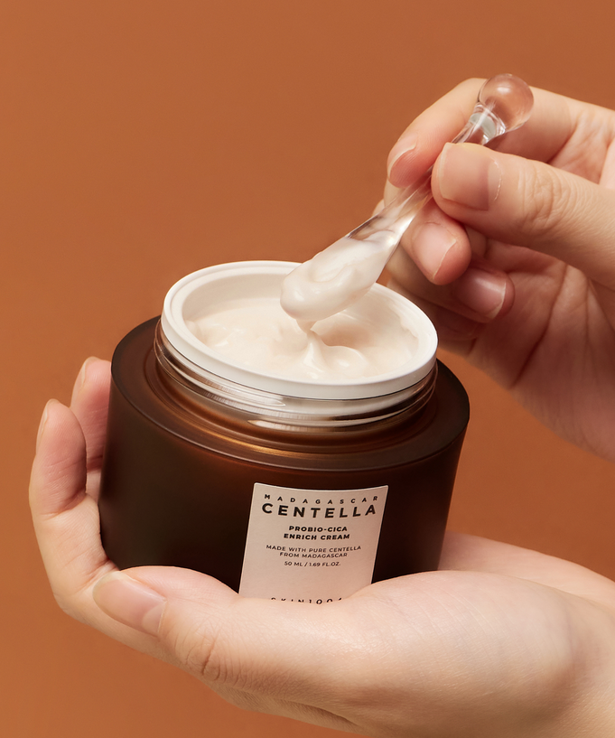 A jar of SKIN1004 Madagascar Centella Probio-cica Enrich Cream 50ml being applied with a spatula against a brown background.