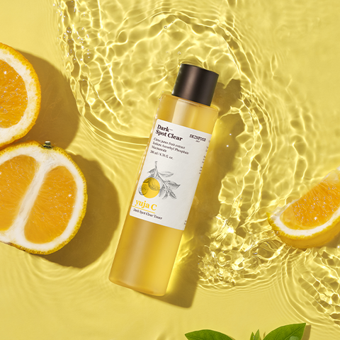 SKINFOOD Yuja C Dark Spot Clear Toner 200ml bottle surrounded by sliced yuja fruit and clear water ripples on a sunny yellow background.