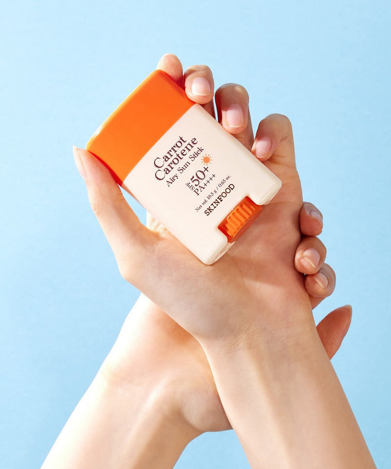 Hands holding SKINFOOD [NEW] Carrot Airy Soft Sun Stick (18.5g) with light blue background.