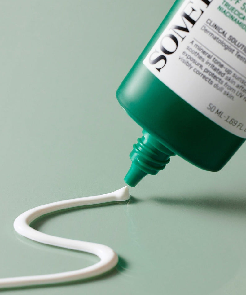 SOME BY MI Truecica Weaponless Calming Tone Up Sun Cream 50ml being dispensed from tube, showcasing texture and color on green surface.