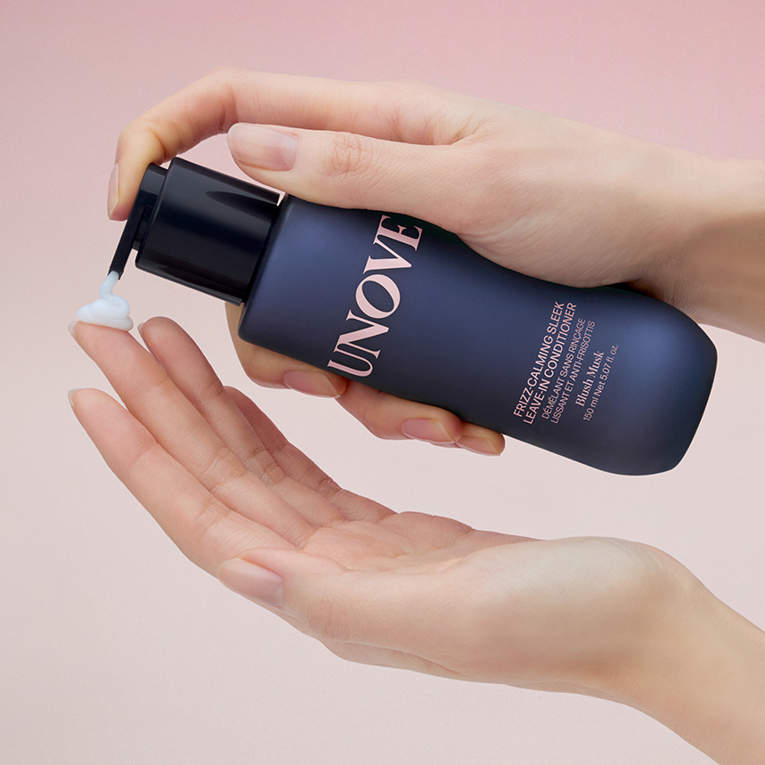 Hands holding a bottle of UNOVE Frizz Calming Sleek Leave-In Conditioner 150ml dispensing product, with a gradient background.