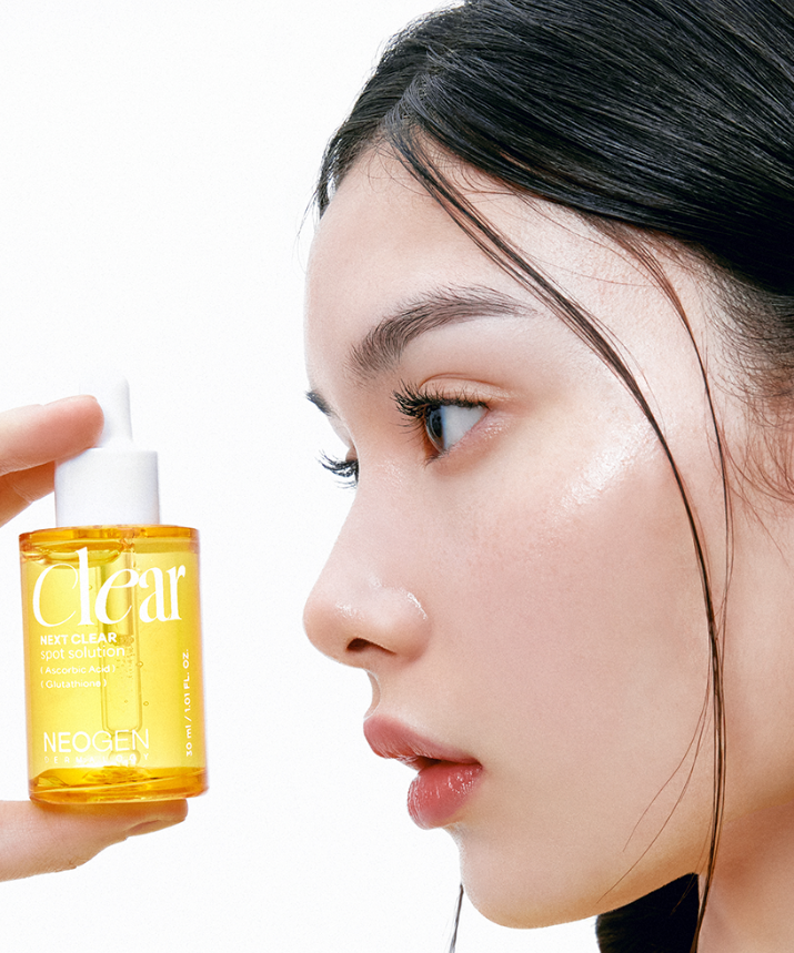Close-up of person holding NEOGEN Next Clear Spot Solution 32ml bottle near face, showcasing skincare product packaging and texture.