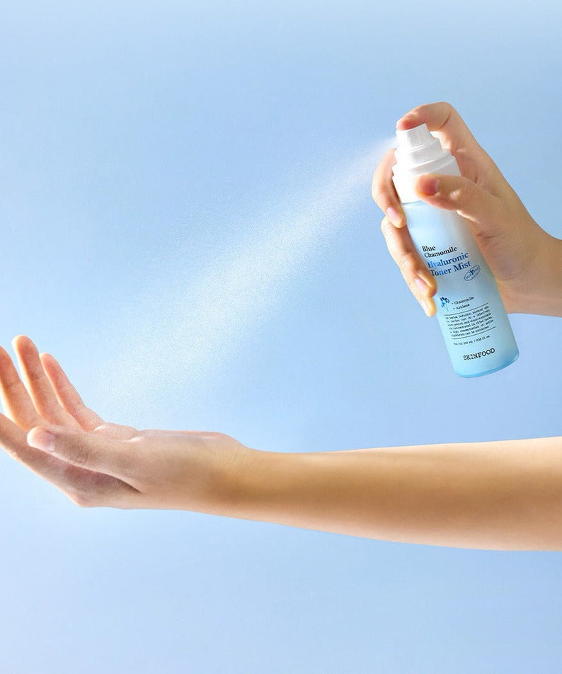 Person spraying SKINFOOD Blue Chamomile Hyaluronic Toner Mist 100ml onto hand against blue background.