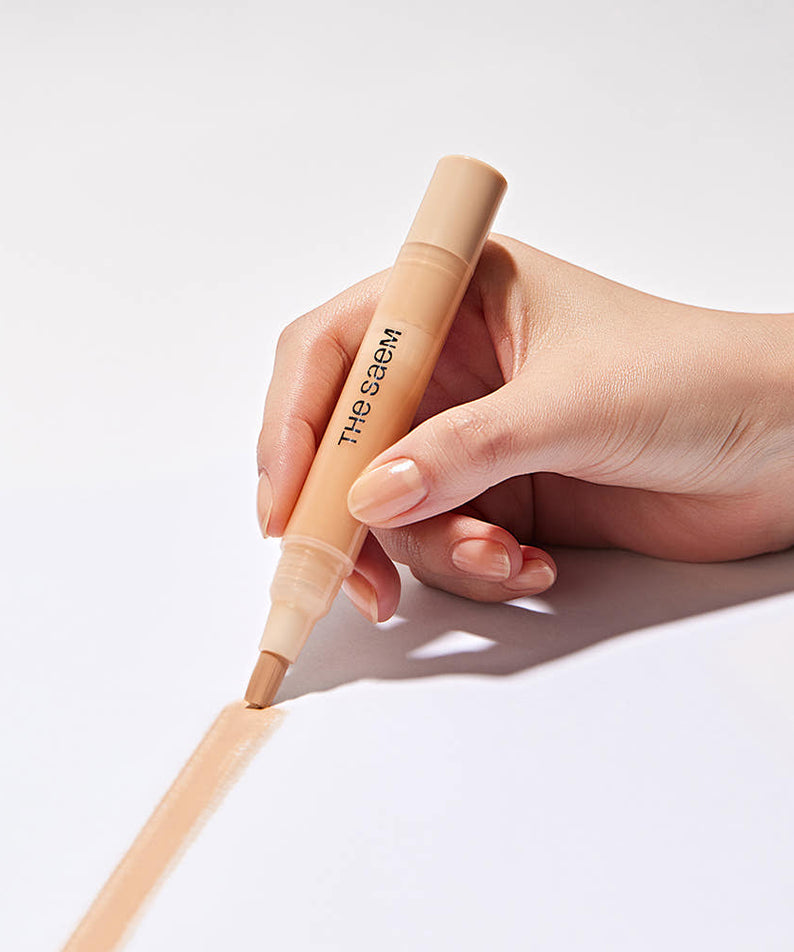 Hand holding THE SAEM Cover Perfection Pen Concealer applying product on white surface