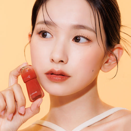 Woman holding THE SAEM Saemmy's Aid Shot Tint against a orange background.