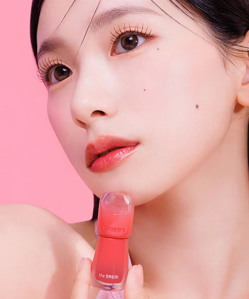 Model holding THE SAEM Saemmy's Aid Shot Tint with vibrant lip color against a pink background.