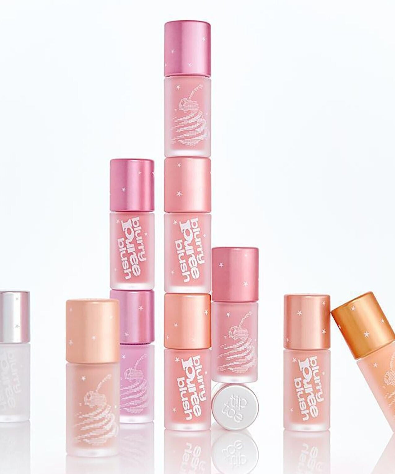 Assorted shades of TIP TOE Blurry Puree Blusher bottles stacked creatively against a white background, showcasing packaging design.