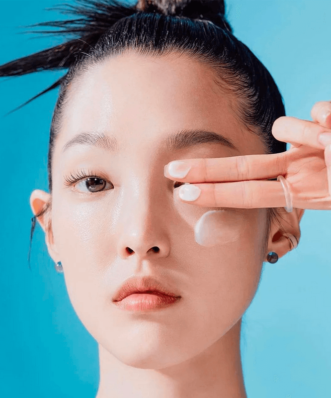 Woman applying TOCOBO Sun Care Mini Duo sunscreen to face, close-up with blue background