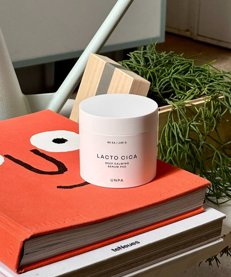 UNPA Lacto Cica Deep Calming Serum Pad 85 Sheets on an orange book beside green plant decor