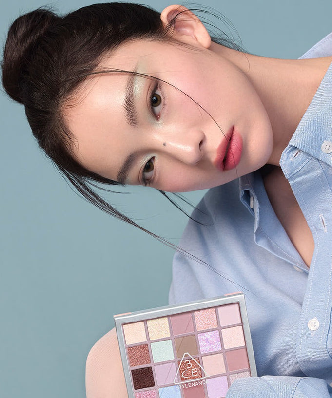 Woman holding 3CE XL Eyeshadow Palette against blue background, showcasing diverse color selection for eye makeup.