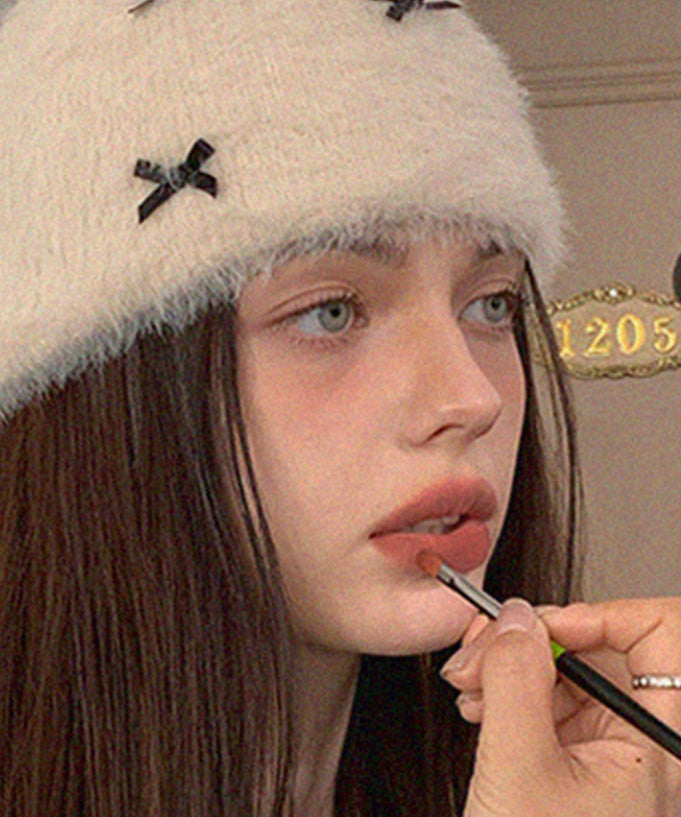 Woman applying AOU Floofy Matte Balm to her lips, wearing a white fuzzy hat with black bows, highlighting the matte finish.