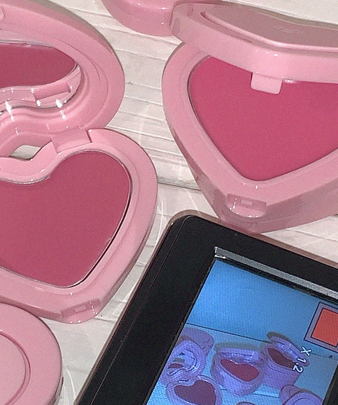 Heart-shaped pink AOU Soft Cream Blush in open compact cases next to a digital display on a wooden surface.