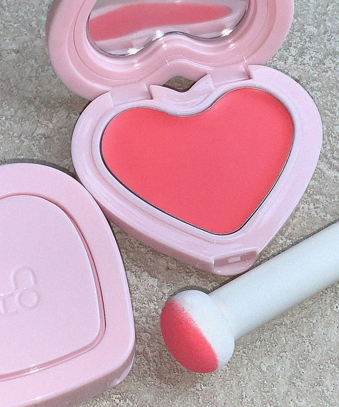AOU Soft Cream Blush in heart-shaped compact with applicator, showcasing its vibrant pink hue on a textured background.