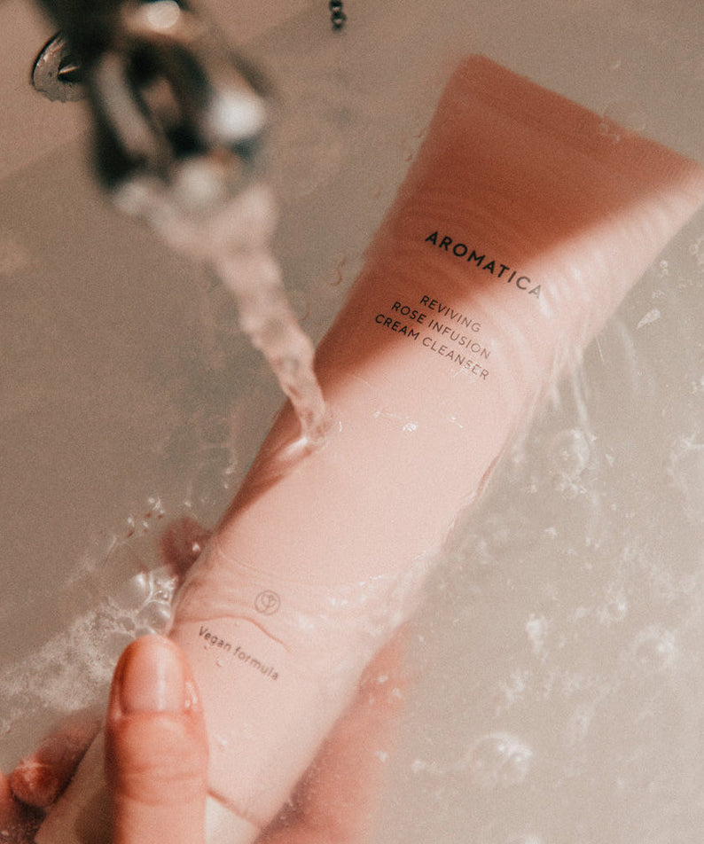 Hand holding AROMATICA Reviving Rose Infusion Cream Cleanser 145ML under running water