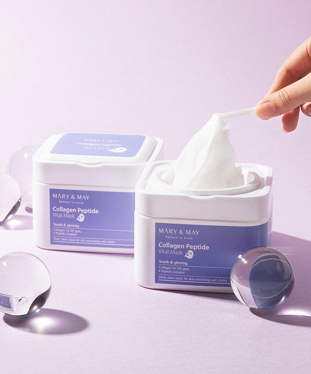 Hand holding tissue from MARY & MAY Collagen Peptide Vital Mask x30 container on a light purple background with glass spheres.