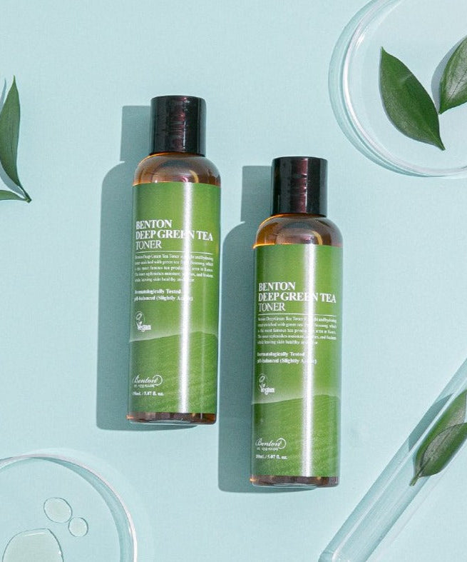 Two bottles of BENTON Deep Green Tea Toner 150ml with fresh green tea leaves on a blue background.