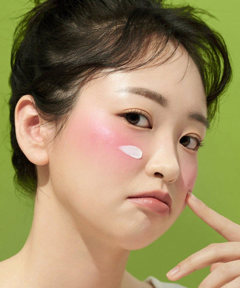 Woman applying FRUDIA Avocado Greenery Relief Sun Cream 50g on her cheek with a green background.