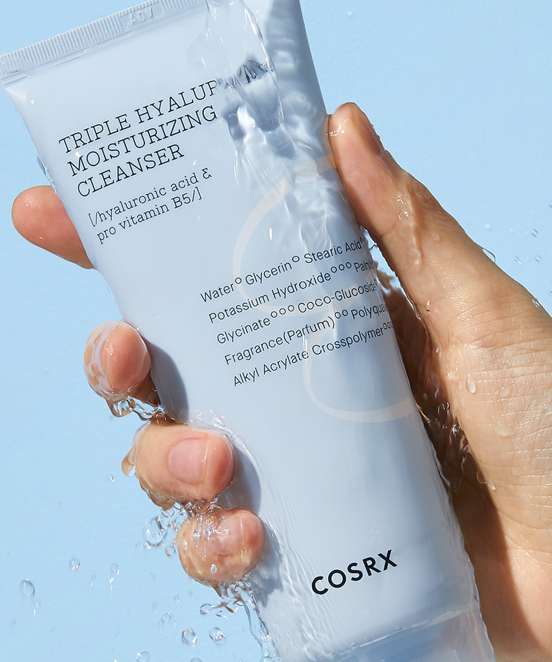 Hand holding COSRX Hydrium triple Hyaluronic Moisturizing Cleanser 150ml with water splash, skincare product display.