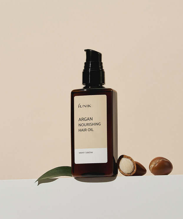 Bottle of iUNIK Argan Nourishing Hair Oil 100ml with argan nuts and a leaf on a neutral background