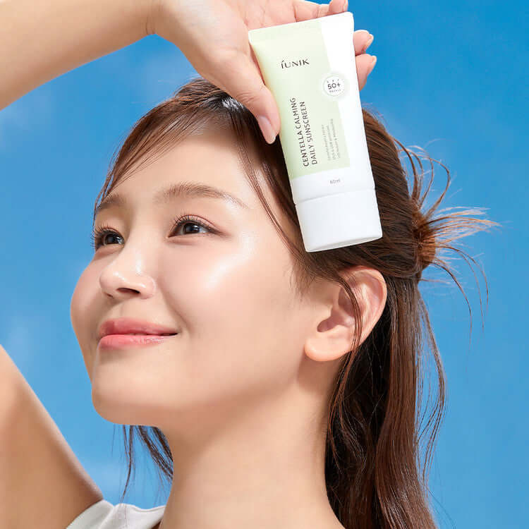 Woman holding iUNIK Centella Calming Daily Sunscreen 60ml against a blue background, smiling with radiant skin.