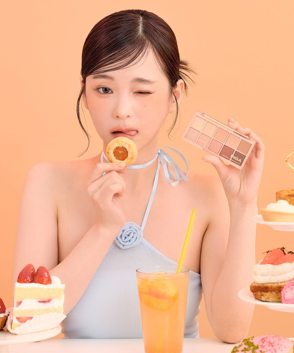 Woman holding INGA Mood Eyes Shadow Palette next to desserts, showcasing elegant makeup and lifestyle scene on an orange background.