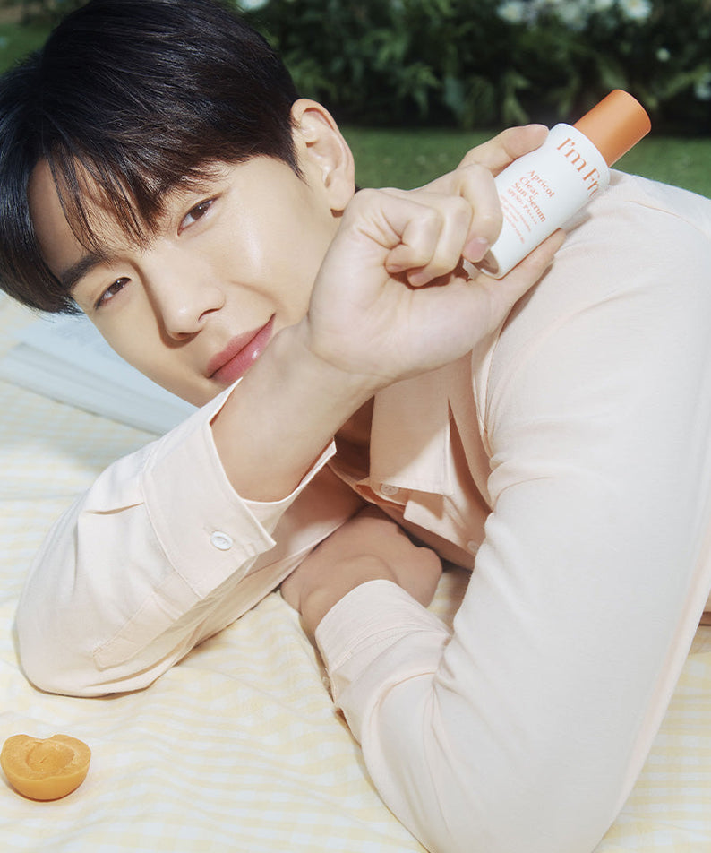 Person holding I'M FROM Apricot Clear Sun Serum 50ml outdoors on pastel blanket with apricots; skincare product displayed.