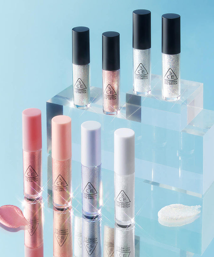 A collection of 3CE Eye Switch liquid eyeshadows in various shades with shimmer effect displayed on acrylic stands against a blue background.