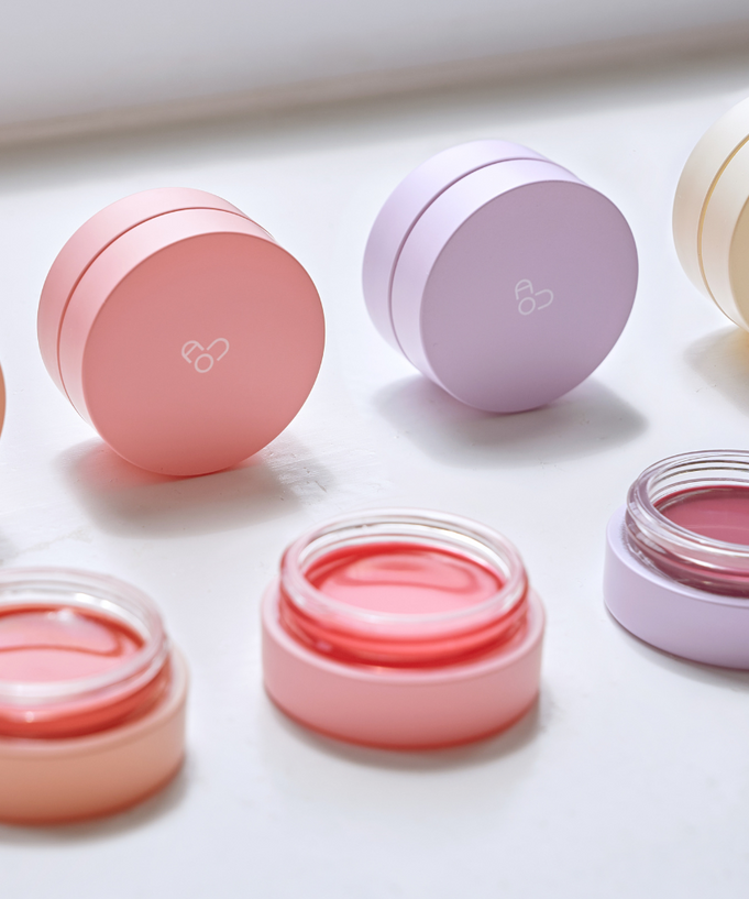 AOU Glowy Tint Balm jars in various pastel colors on white surface, showcasing the product's attractive packaging and shades.