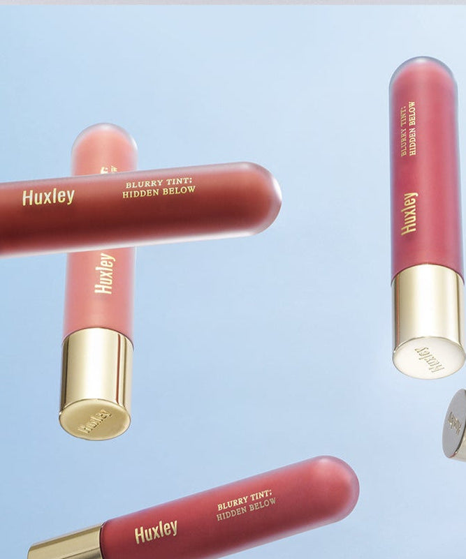 HUXLEY Blurry Tint Hidden Below cosmetic tubes floating in front of a blue background, showcasing elegant design and color.