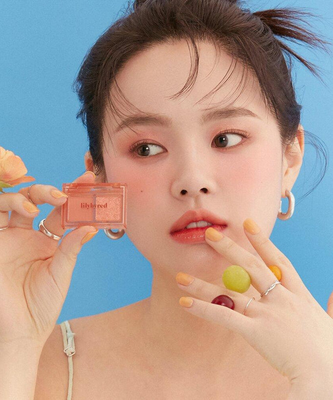 Woman showcasing LILYBYRED Little Bitty Moment Shadow against a blue background with orange flower and vibrant nail polish.