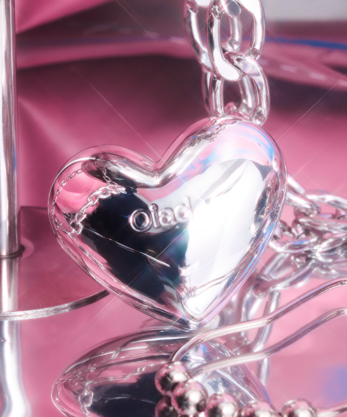Silver heart-shaped pendant with 'oiad' engraving on a chain, set against a pink reflective background.