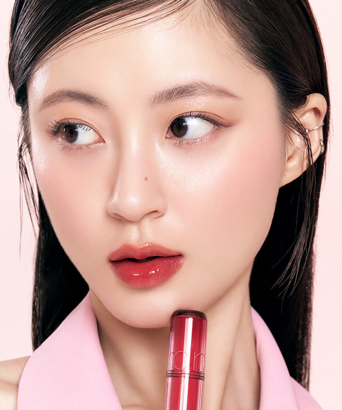 Model applying vibrant ROMAND The Juicy Lasting Tint to lips, showcasing glossy finish and intense color on pale pink background.