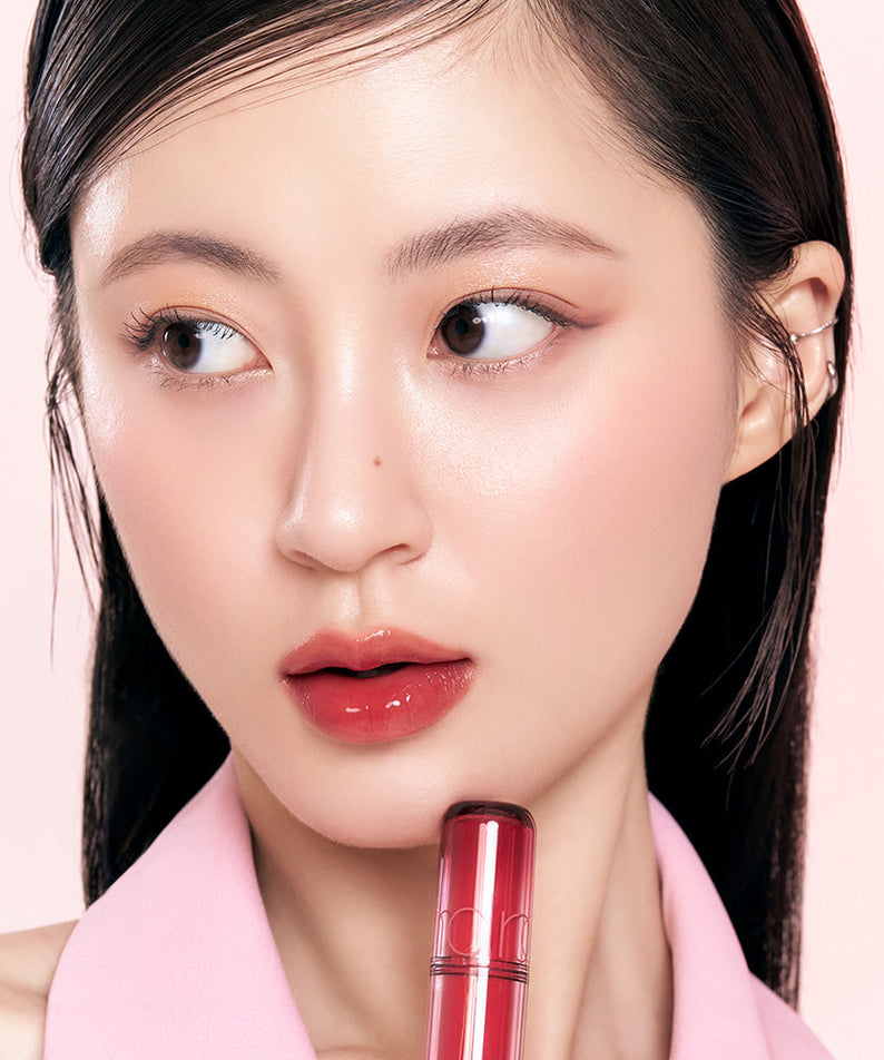 Model applying vibrant ROMAND The Juicy Lasting Tint to lips, showcasing glossy finish and intense color on pale pink background.