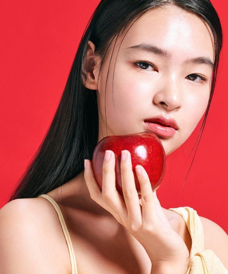 Woman holding an apple next to her face in front of a red background, promoting TOCOBO Apple Dewy Fit Cushion.