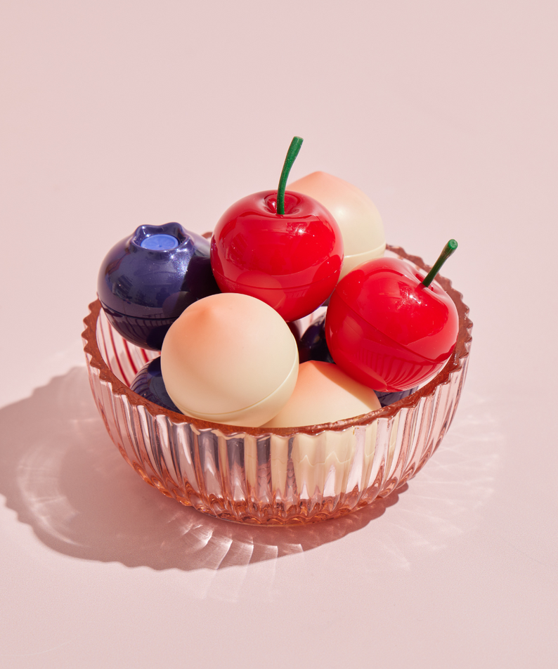 TONY MOLY MIN Fruit Lip Balm in a glass bowl, showcasing colorful fruit-themed packaging on a soft pink background.